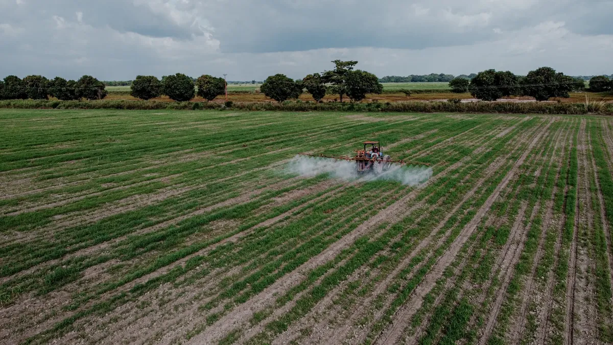 Aerial shot of a tractor spraying a lush green field with pesticides under a cloudy sky.